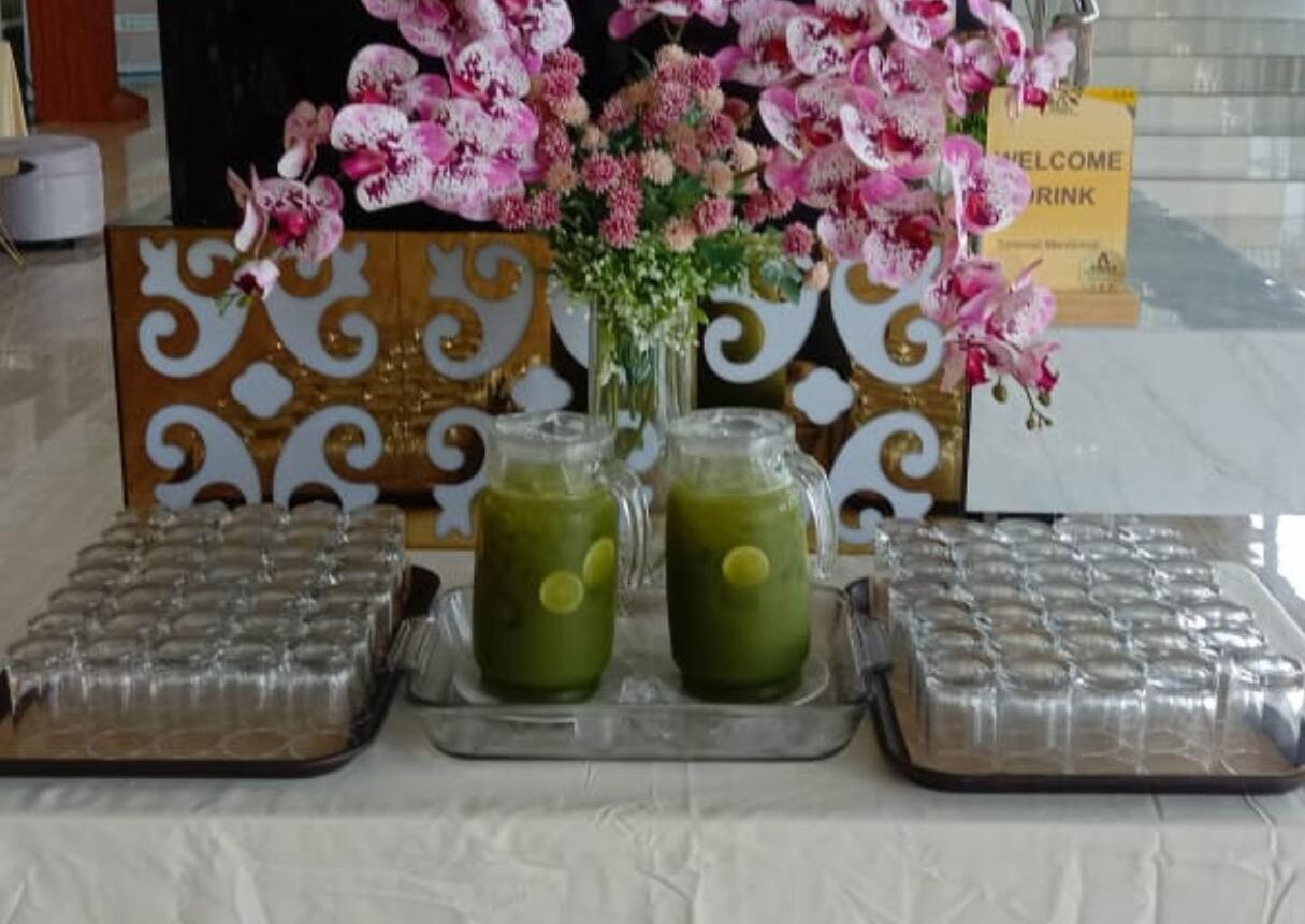 Una mesa con dos bebidas verdes, un florero con flores rosadas y un letrero de bienvenida.