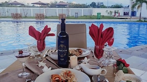 A dining table by a pool with plates of food, glasses, and a bottle of Mascalchi Port wine. - Aimas Hotel & Convention Centre (Sorong)