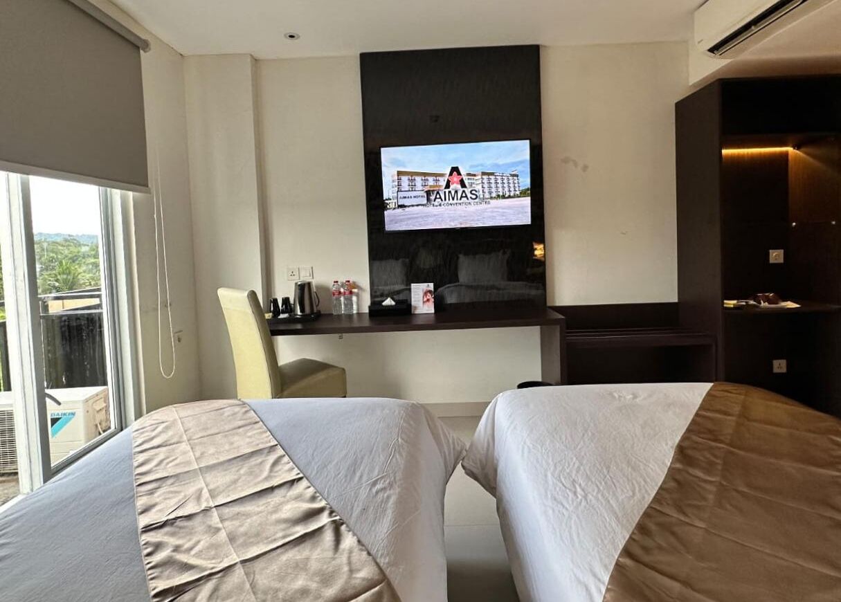 A hotel room with two beds, a TV mounted on the wall, a desk with a chair, and a view of a building outside.
