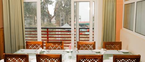 A dining area with a table set for six, wooden chairs with intricate back designs, and a view through a large window to the outside.