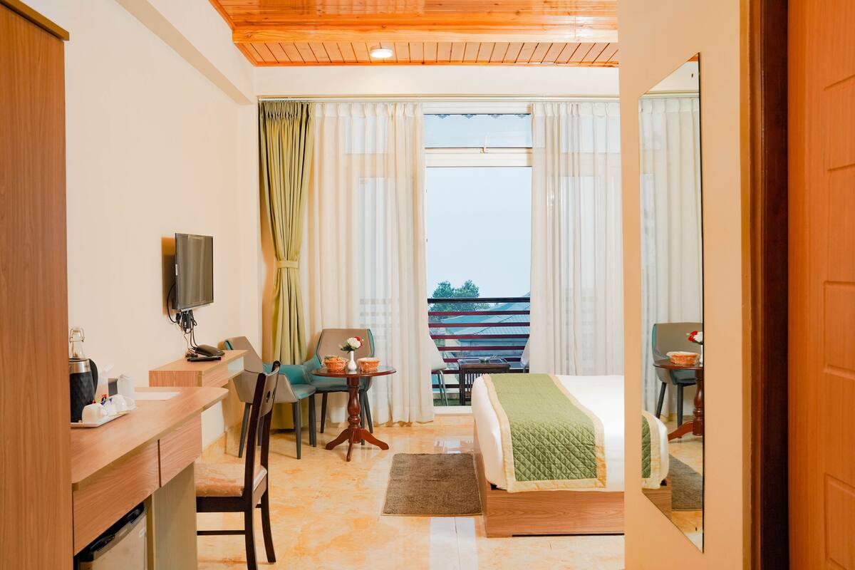 A hotel room with a bed, a desk with a chair, a small table with a chair, a TV, and a balcony with curtains.