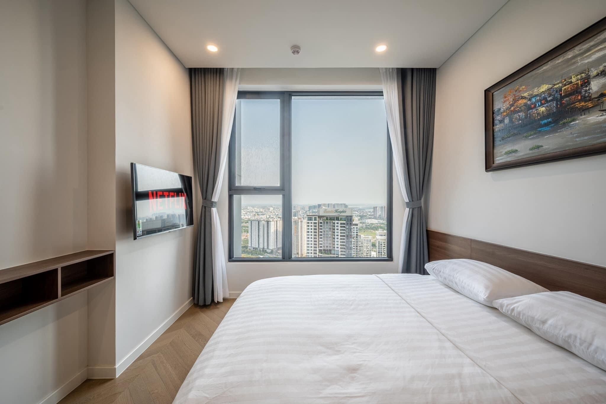 A modern hotel room with a large bed, a TV mounted on the wall, and a city view through the window.