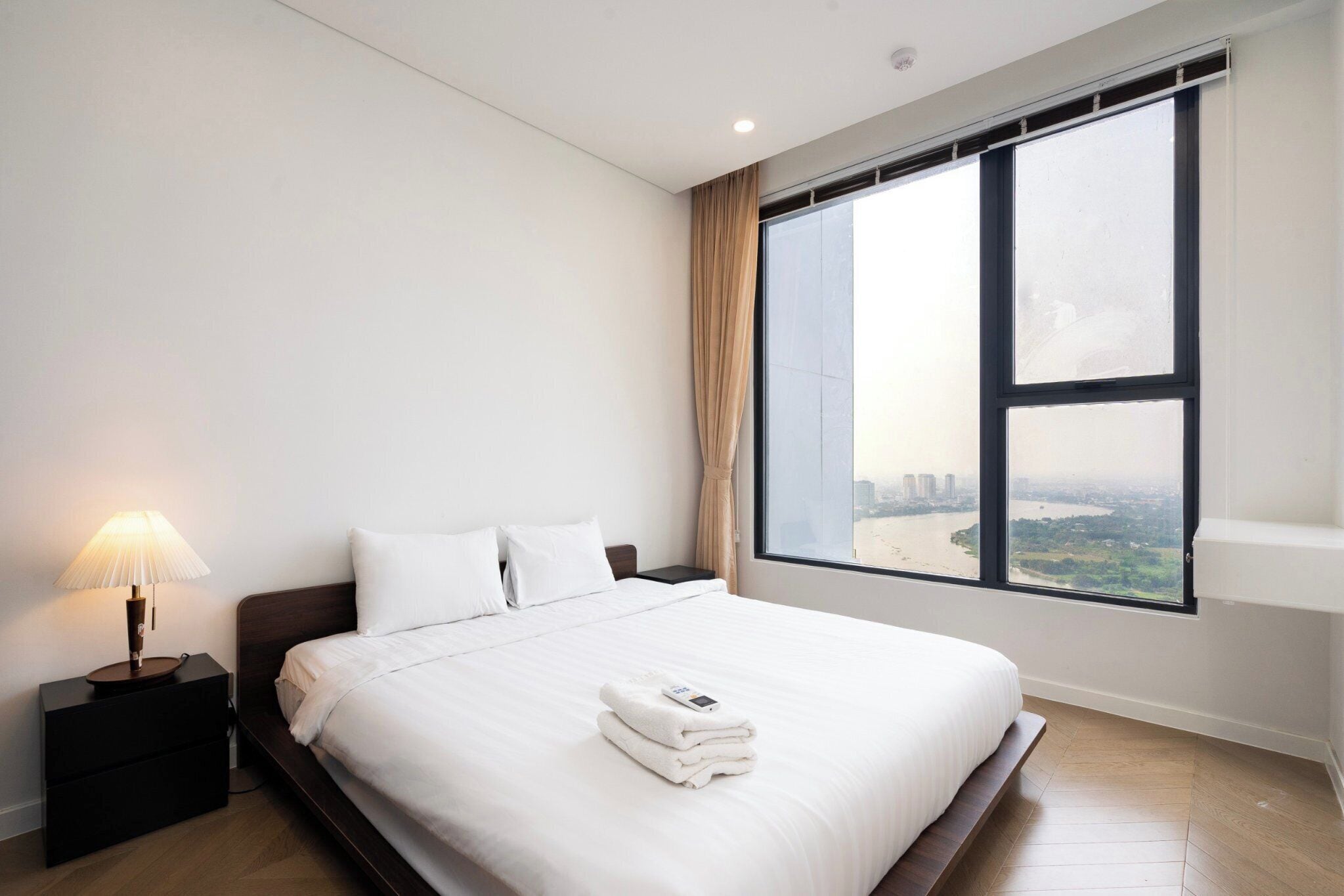 Een hotelkamer met een groot bed, een nachtkastje met lamp en uitzicht op een stadsbeeld door een groot raam.