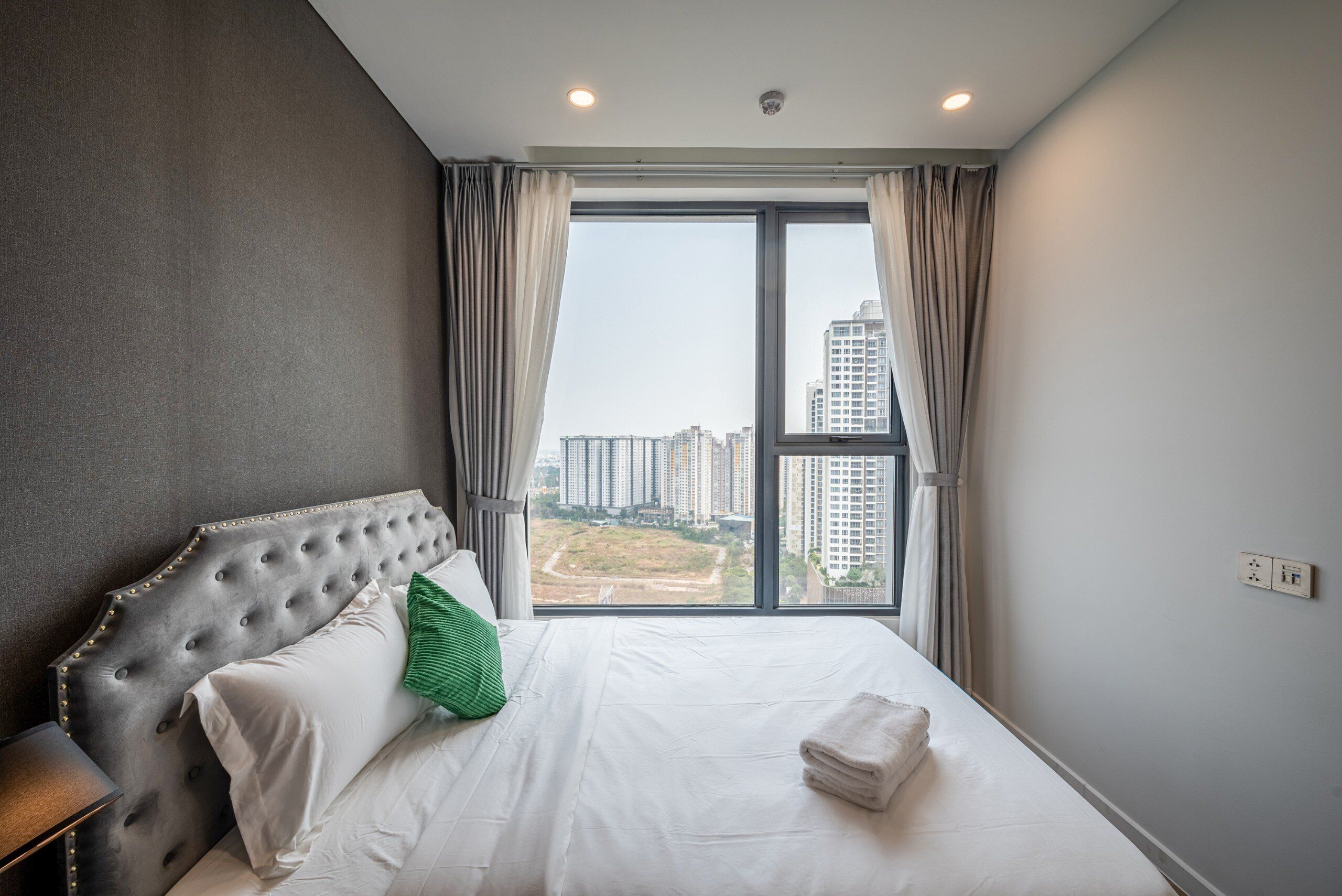 Habitación de hotel con una cama grande, cabecera capitonada, vista a edificios altos y ventana con cortinas.