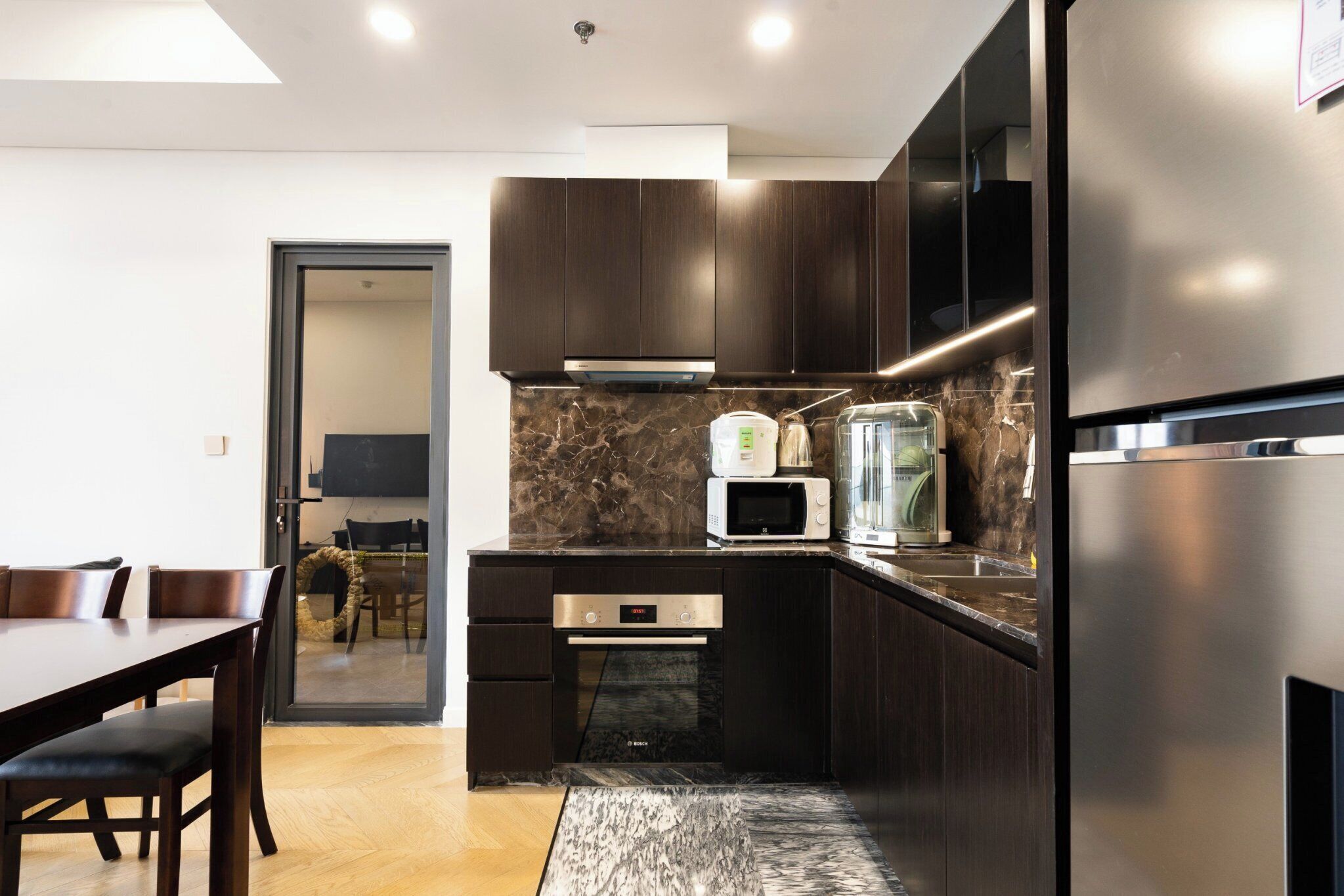 A modern kitchen with dark wood cabinets, a stainless steel refrigerator, a built-in oven, and a marble backsplash.