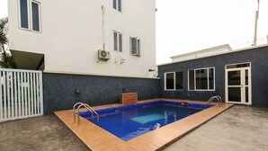 Ein rechteckiger Pool mit Holzterrasse, umgeben von einem weißen Gebäude mit Fenstern und einer grauen Mauer.