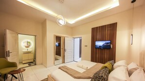 Ein modernes Schlafzimmer mit Bett, Fernseher, grünem Sessel, kleinem Tisch und einem Badezimmer mit Spiegel und Waschbecken.