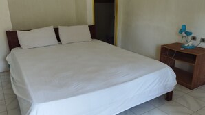 Habitación con una cama individual, cabecera de madera, ropa de cama blanca, una mesita de noche de madera con una lámpara azul y una puerta que da a una habitación oscura.