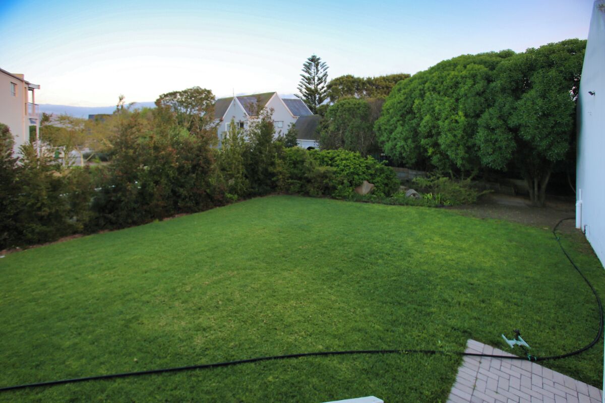 A well-maintained grassy backyard with a paved area, surrounded by trees and a white building.