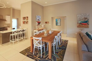 A modern kitchen with a central island, white bar stools, a wooden dining table with white chairs, and artwork on the walls.