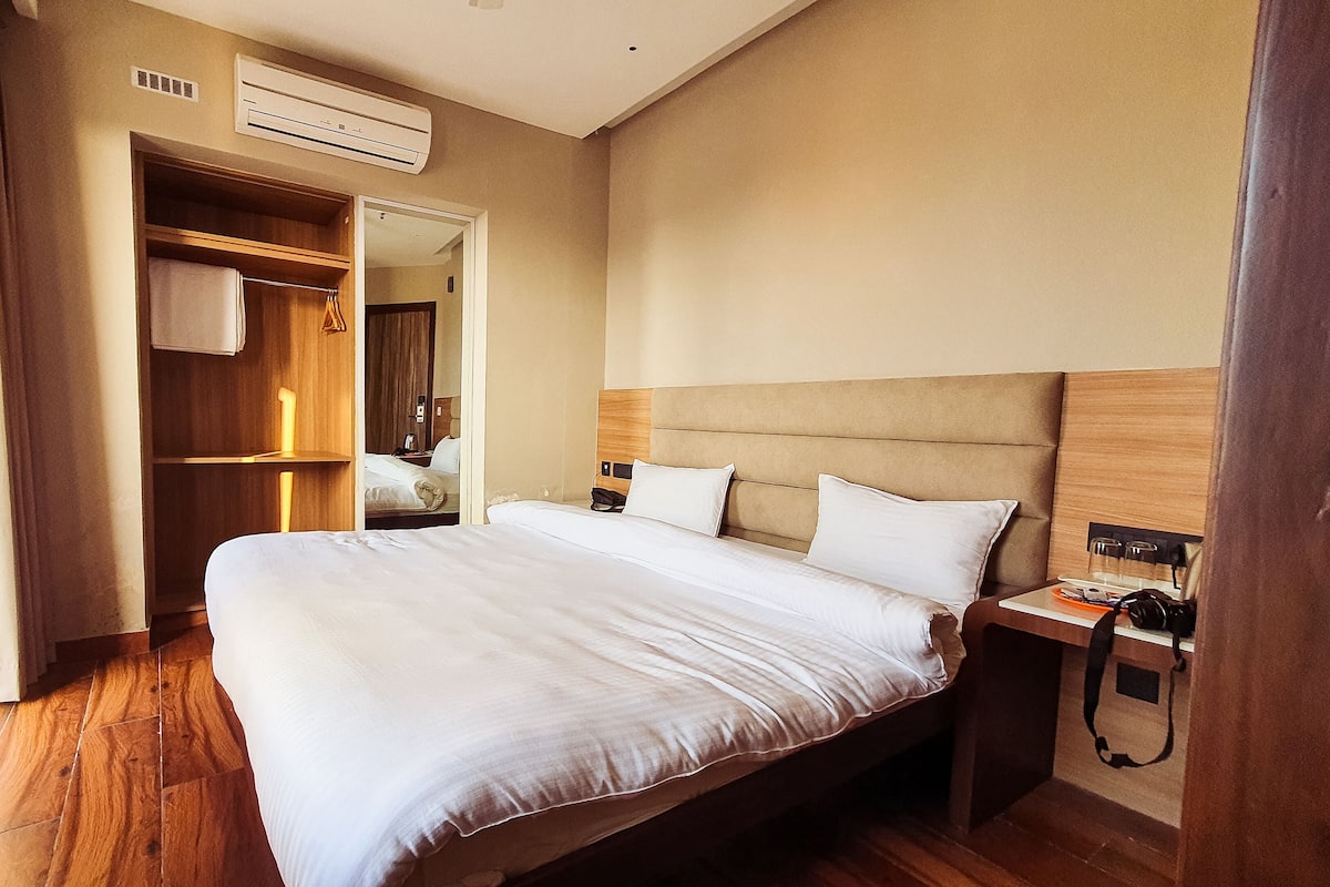 Una habitación de hotel con una cama grande, suelo de madera, un armario y una mesita de noche con lámpara.