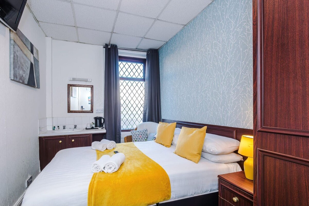A hotel room with a bed, a bedside table with a lamp, a window with curtains, a bathroom with a mirror and sink, and a wall with a patterned design.