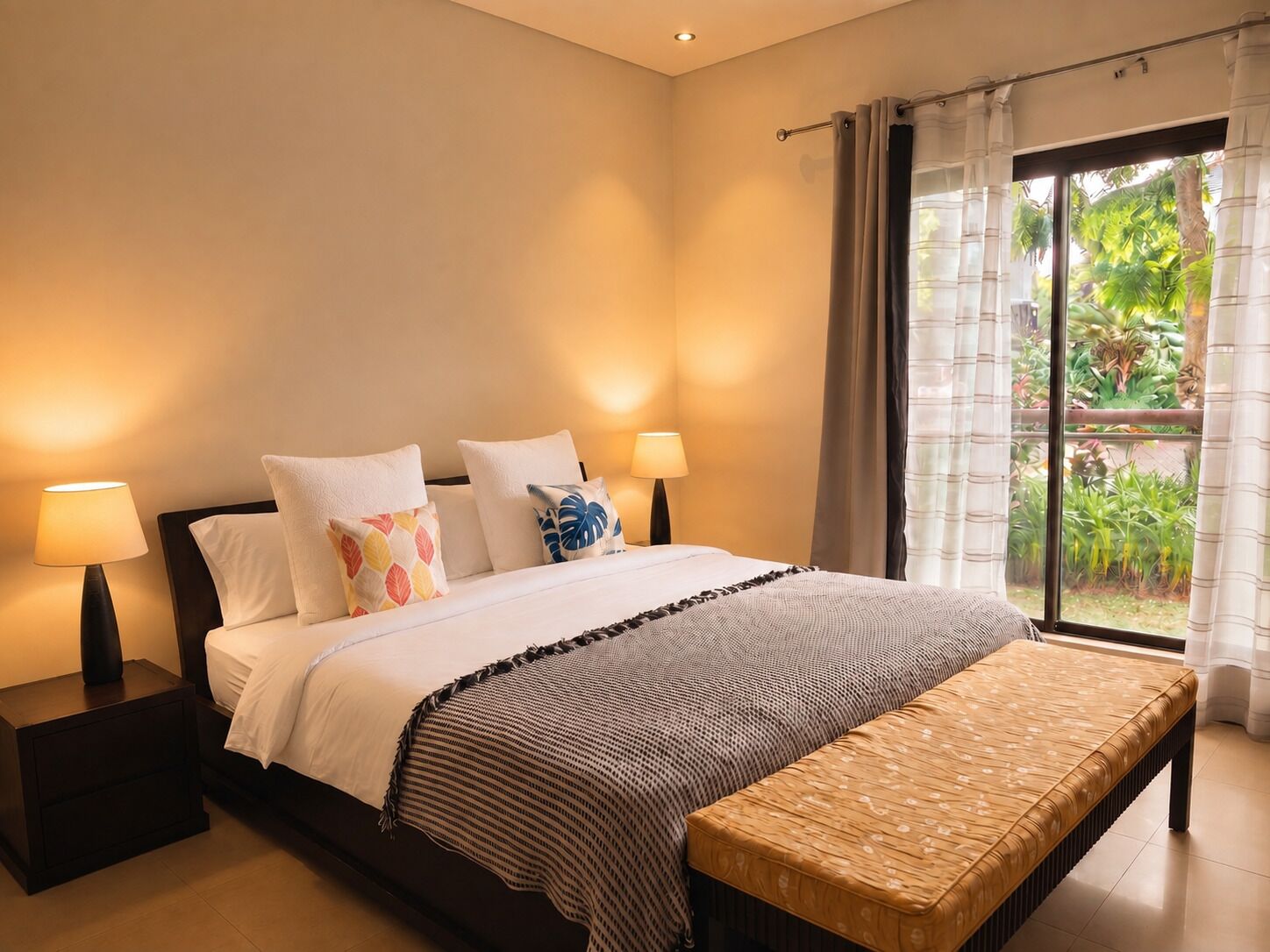 A hotel room with a bed, bedside lamps, a bench, and a view of greenery through the window.