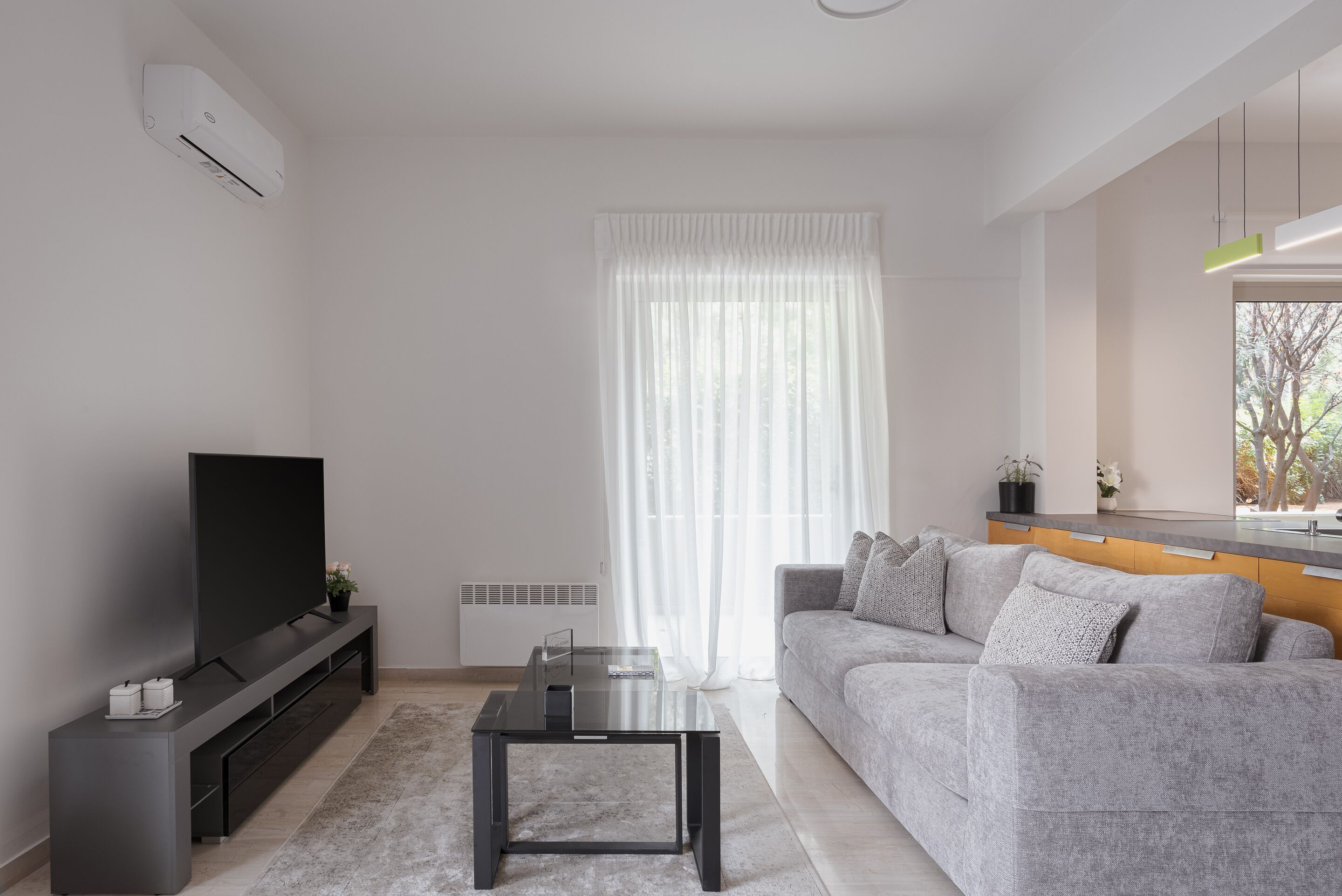 Een moderne woonkamer met een flatscreen televisie, een glazen salontafel, een grijze bank en een houten dressoir.