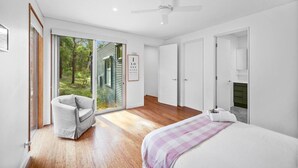 A modern bedroom with a large sliding glass door leading to a garden, a bed with a checkered blanket, a chair, and a ceiling fan. - Aireys Bush Retreat - Spacious home with huge deck (Aireys Inlet)