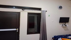A room with a dark wooden door, a window with a curtain, a wall-mounted TV, and a bed with a star-patterned bedspread. - Hotel O Ayna Kost Syariah (Singaraja)