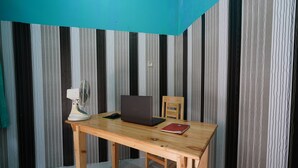 A wooden desk with a laptop, a fan, and a red folder in a room with striped wallpaper. - Hotel O Ayna Kost Syariah (Singaraja)