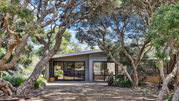 Una casa moderna con un amplio ventanal, rodeada de árboles y un camino de entrada de grava.