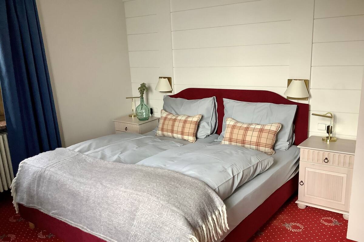 A neatly made bed with a red headboard, two bedside tables with lamps, and a red patterned carpet.