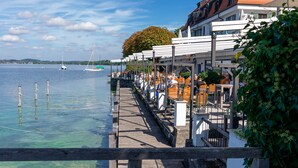 A waterfront restaurant with outdoor seating, a wooden deck, and a view of the lake and sailboats. - Strandhotel Berg (Berg)