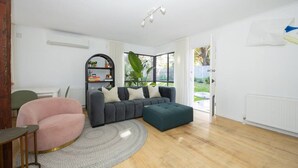 A modern living room with a grey sofa, a pink armchair, and a green ottoman. There is a bookshelf with decorative items and a large window with a view of the outdoors.