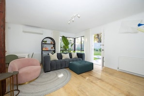 A modern living room with a grey sofa, a pink armchair, and a green ottoman. There is a bookshelf with decorative items and a large window with a view of the outdoors.