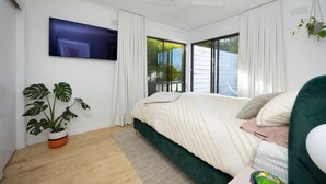 A modern bedroom with a large bed, a ceiling fan, a TV mounted on the wall, and a large window with curtains.