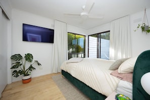 A modern bedroom with a large bed, a ceiling fan, a TV mounted on the wall, and a large window with curtains.