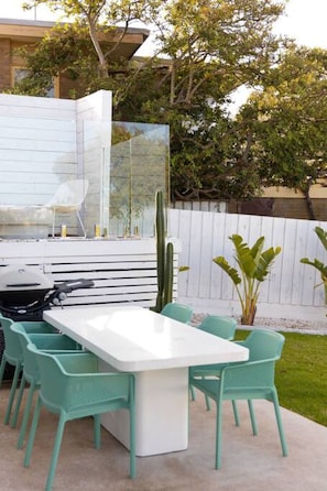 An outdoor patio with a white table and turquoise chairs, surrounded by greenery and a cactus.