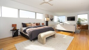 A modern bedroom with a large bed, a white sofa, a ceiling fan, and a TV mounted on the wall.