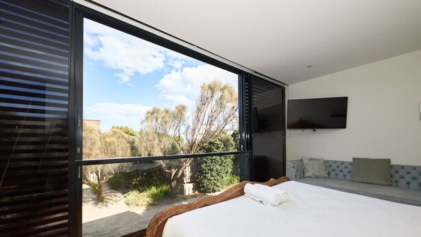 A modern bedroom with a large window, a bed, a flat-screen TV, and a view of trees and a building outside.
