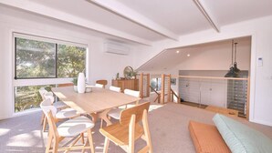 Ruime kamer met een grote houten eettafel, witte stoelen, een beklede bank en uitzicht op groen.