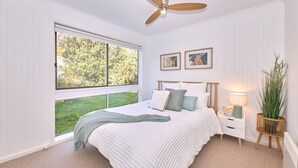 Een slaapkamer met een groot raam, een bed met een witte deken, een houten hoofdbord, een nachtkastje met een lamp en een plafondventilator.
