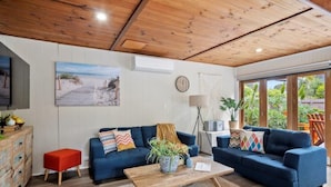 Una sala de estar acogedora con un sofá azul, una mesa de centro de madera y un taburete rojo. Se ve una ventana grande con vistas a la vegetación.