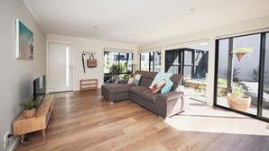 A spacious living room with a sectional sofa, wooden flooring, a TV, and a sliding glass door leading to an outdoor area with plants.