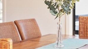 A dining table with a clear vase and eucalyptus branches, a clock with 'BEACH' written on it, and wicker chairs.