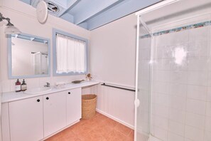 Une salle de bain avec un lavabo blanc, deux miroirs, une fenêtre avec des rideaux blancs, une douche avec une porte en verre et un panier en osier posé sur le sol.