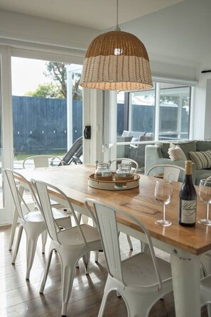 A dining area with a wooden table, white chairs, a wicker pendant light, and a view of a backyard through large sliding glass doors. - Coastal Family Lodge (Torquay)