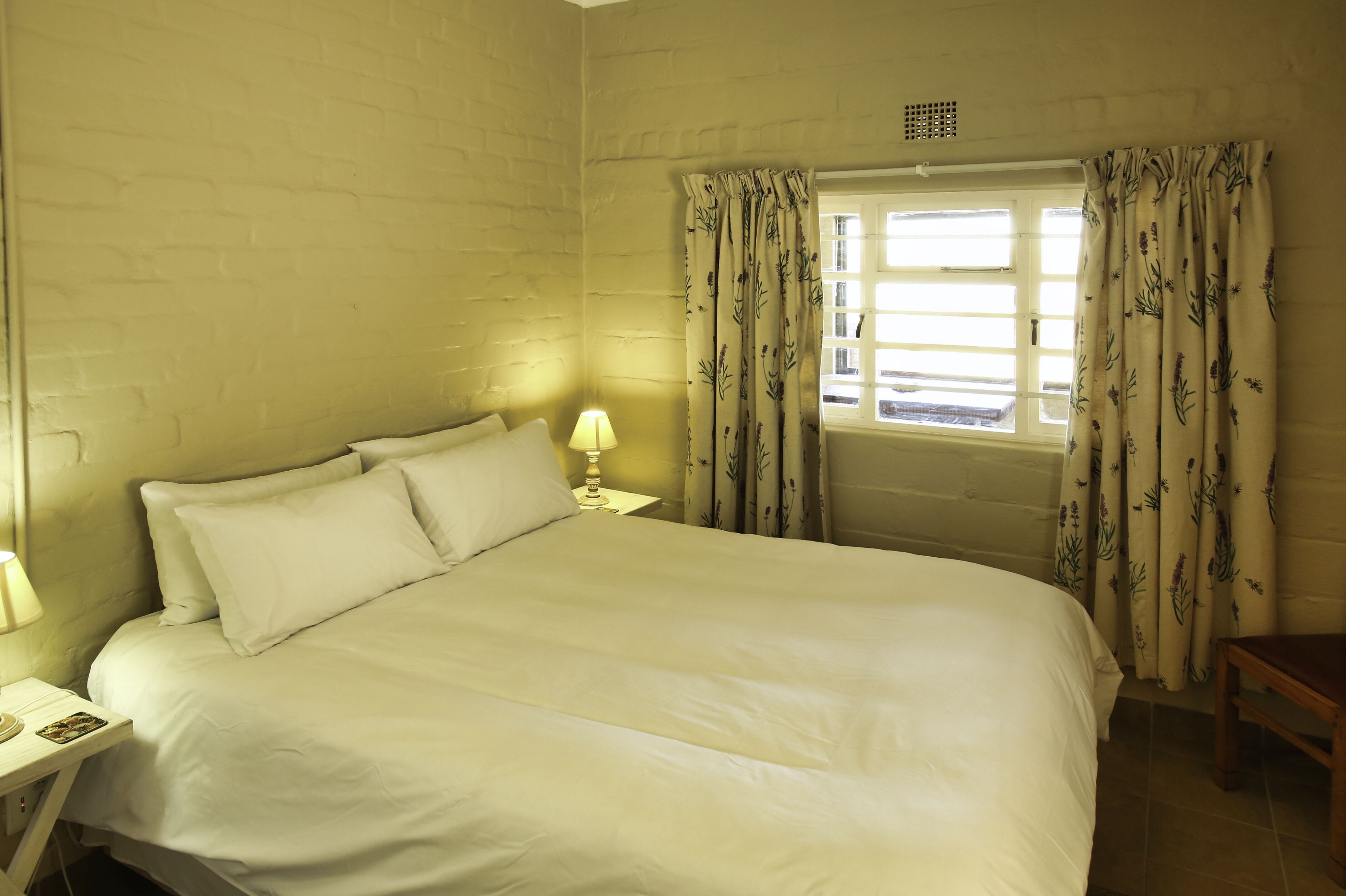 A bedroom with a large bed, a nightstand with a lamp, a window with floral curtains, and a wooden bench.
