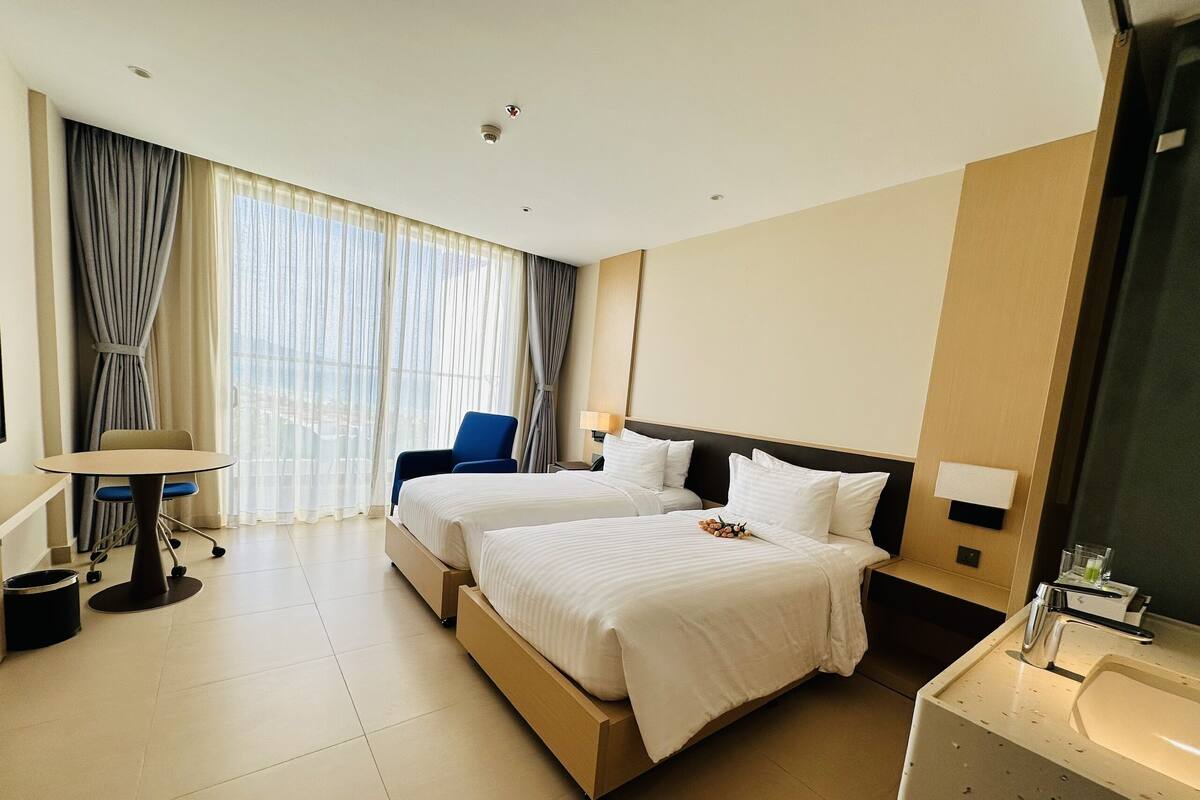 A hotel room with two beds, a desk, a chair, a desk lamp, a nightstand, and a bathroom with a sink and mirror.