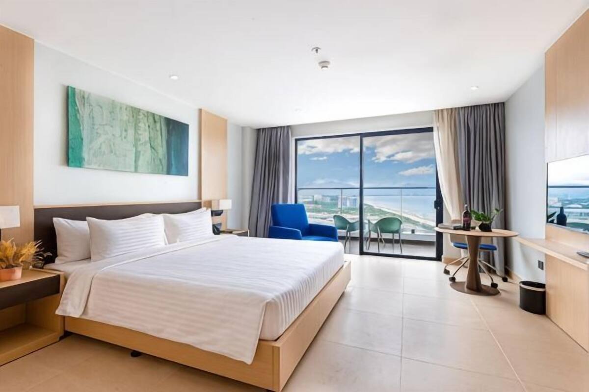 A modern hotel room with a large bed, a desk, a chair, and a balcony with a city view.