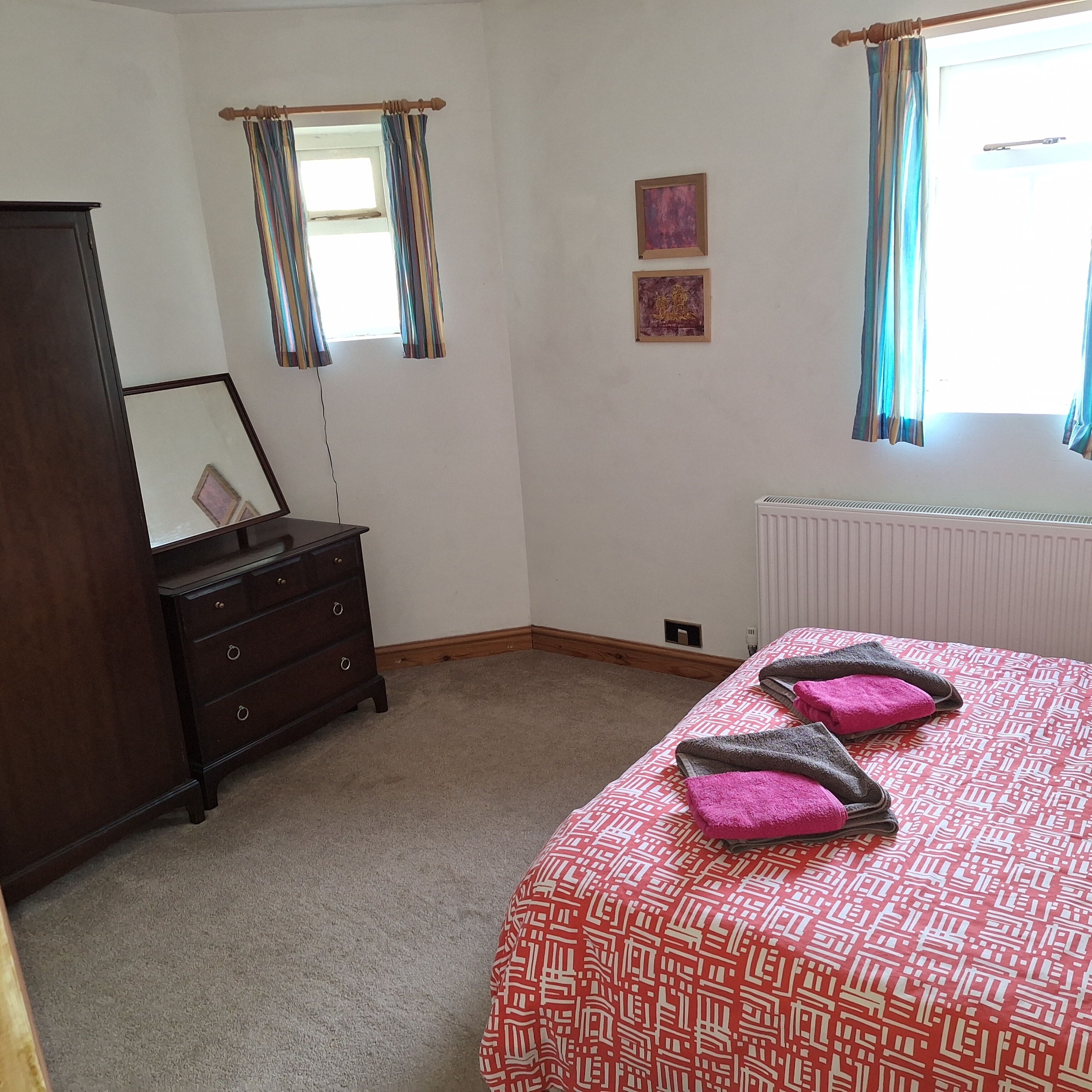 A bedroom with a bed, a dresser, a radiator, and two windows with curtains.