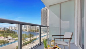 A balcony with a view of a cityscape, a chair, and a table with a bottle and a glass.
