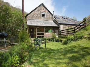 A stone building with a sloped roof, a wooden porch, and a fenced garden area. - Watermill Cottages (Dartmouth)