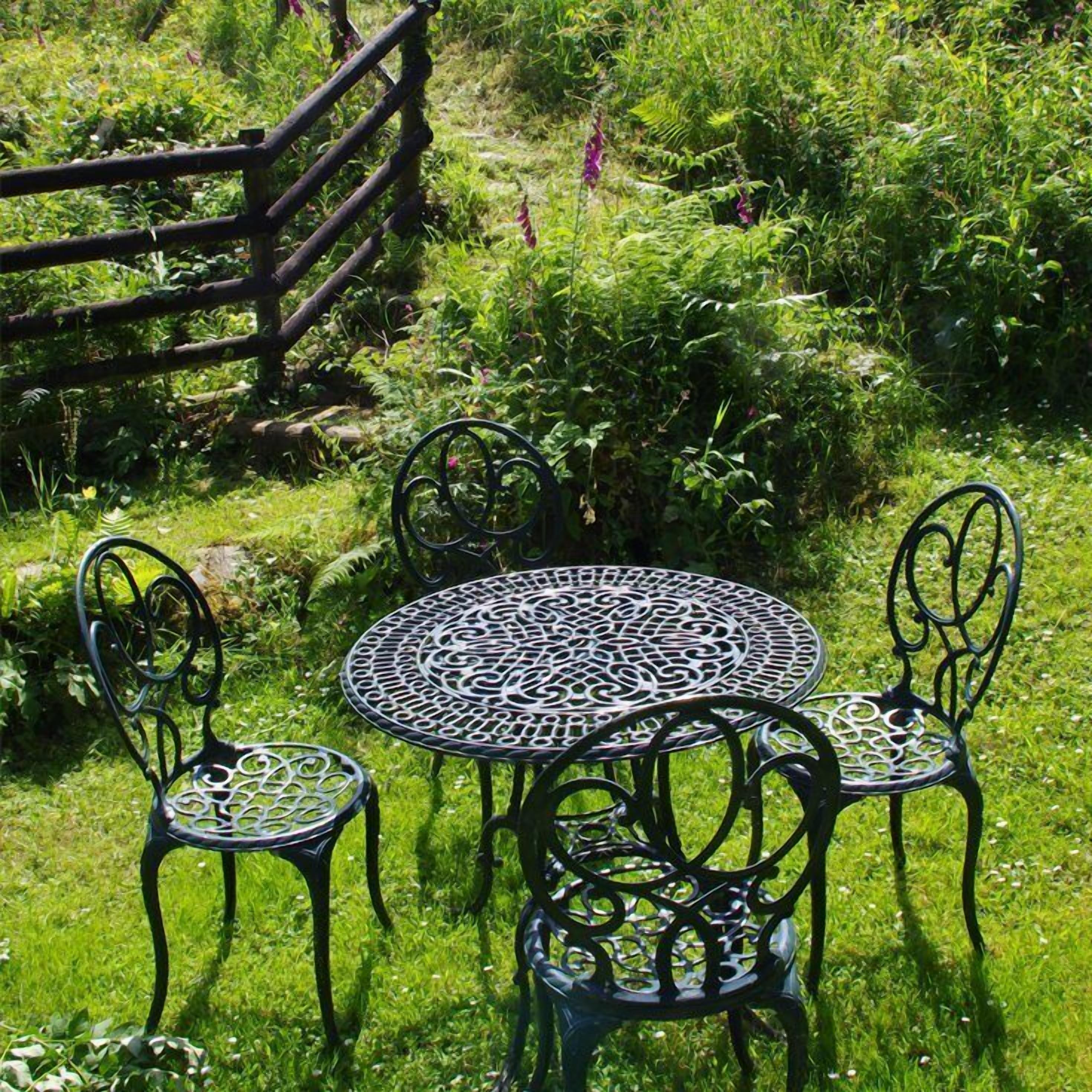 Een ronde metalen tafel met ingewikkelde patronen, omringd door vier metalen stoelen met vergelijkbare designs, geplaatst op een gazon met een houten omheining op de achtergrond.