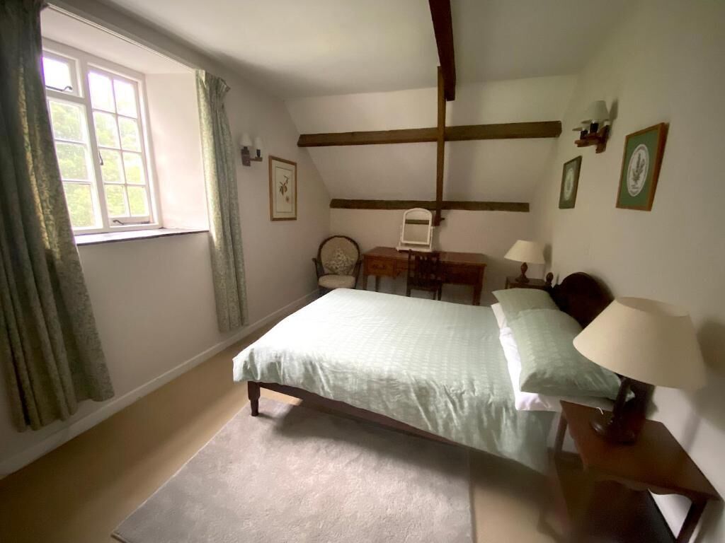 A bedroom with a bed, a nightstand, a chair, a window with curtains, and a wooden ceiling beam.