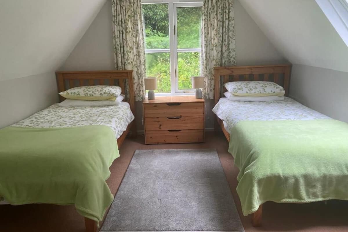 Una habitación pequeña y acogedora con dos camas individuales, un aparador de madera, una ventana con cortinas y vistas a zonas verdes.