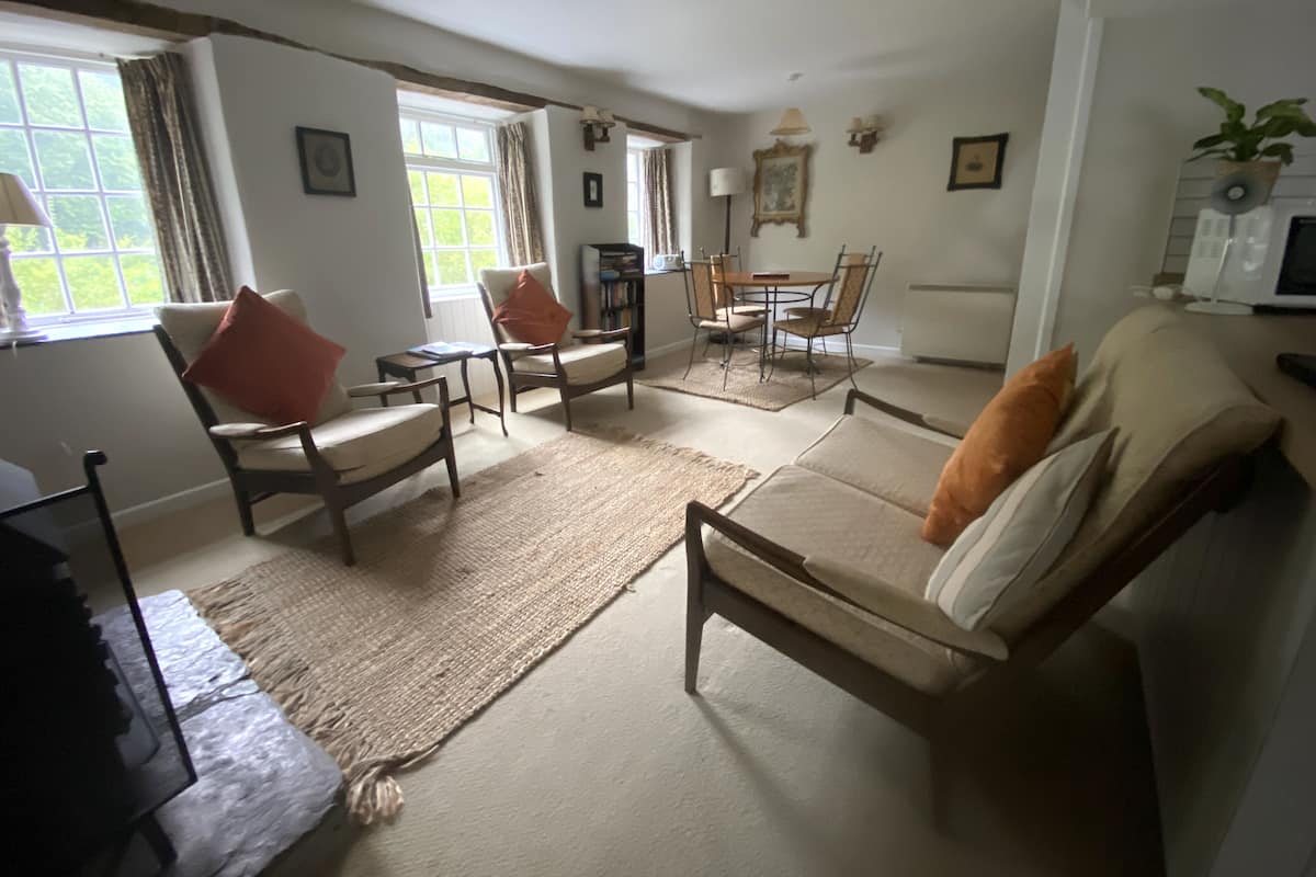 Un salón con sofá, sillones, una mesa de centro y una mesa de comedor con sillas. Hay una estantería, una chimenea y una ventana con cortinas.