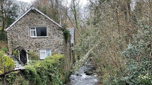 A stone house with a small window and a door, surrounded by dense vegetation and a narrow stream. - Watermill Cottages (Dartmouth)