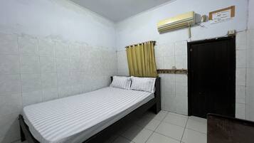 Quarto pequeno com uma cama de solteiro, paredes de azulejos brancos, uma porta preta e uma janela com cortina.